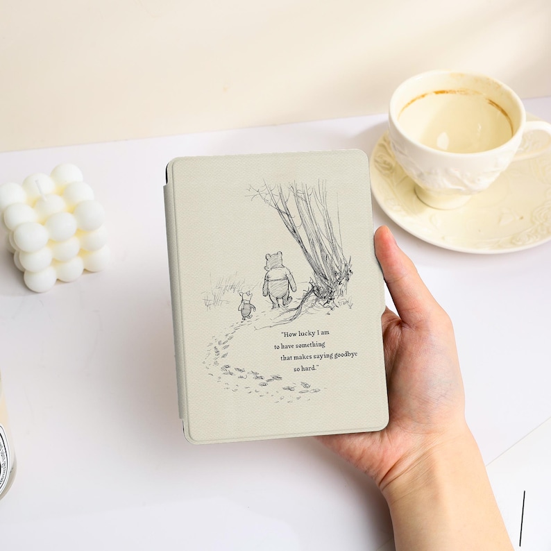 May include: A light beige e-reader cover featuring a black ink illustration of Winnie the Pooh and Piglet walking away. The text reads, "How lucky I am to have something that makes saying goodbye so hard." The cover has a smooth, matte finish.