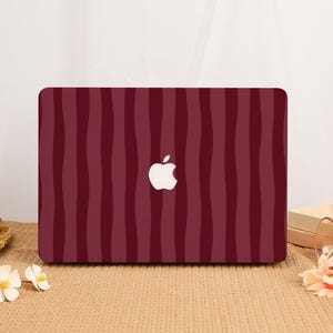 Peut inclure: Une coque d'ordinateur portable arborant un motif de rayures verticales ondulées dans des tons bordeaux profond et rouge foncé. La coque présente une découpe centrale laissant apparaître le logo Apple blanc. Le design alterne des nuances de rouge.