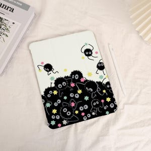 Puede incluir: Funda blanca para tableta con una ilustración en blanco y negro de duendes de hollín y estrellas de colores. La funda tiene una cubierta plegable y se ve un lápiz óptico blanco.
