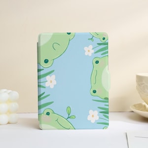 May include: A light blue tablet case featuring a repeating pattern of smiling green frogs, white flowers, and green leaves. The case has a smooth finish and a rectangular shape, designed to protect a tablet.
