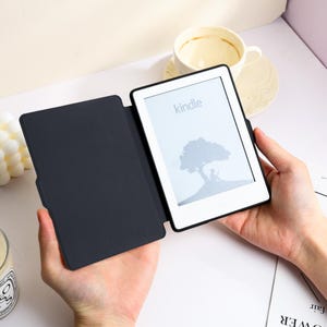 May include: A black e-reader device with a protective cover open to reveal a white screen displaying the word kindle and a graphic of a person sitting under a tree. The device has a slim black frame and a matte black interior cover surface.
