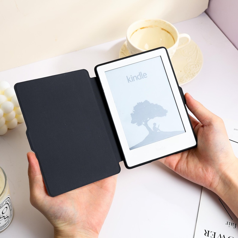 May include: A black e-reader device with a protective cover open to reveal a white screen displaying the word kindle and a graphic of a person sitting under a tree. The device has a slim black frame and a matte black interior cover surface.