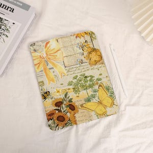 Könnte beinhalten: Eine dekorative Tablet-Hülle mit einem Collage-Design mit Sonnenblumen, einer gelben Schleife, einer Biene und einem Schmetterling. Die Hülle hat eine Vintage-Ästhetik mit Schrift- und floralen Elementen, und ein weißer Stift liegt daneben.