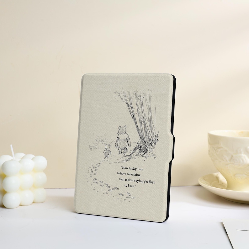 May include: A cream-coloured e-reader case featuring a black line-art illustration of Winnie the Pooh and Piglet walking away. The text reads, "How lucky I am to have something that makes saying goodbye so hard." The case has a black edge and frame.