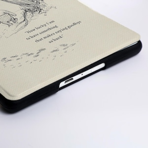 May include: A cream-coloured tablet case with a black frame featuring a line-art illustration of a character and the text, "How lucky I am to have something that makes saying goodbye so hard." The case has a textured surface and a visible charging port.