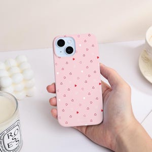 Puede incluir: Una funda de teléfono rosa con un patrón de pequeños corazones rojos y blancos. La funda se sostiene en una mano y tiene un recorte para la cámara. La funda del teléfono es de color liso con un patrón de corazones repetidos.