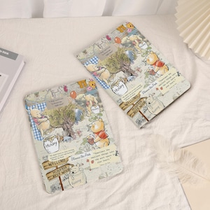 May include: Two rectangular cases featuring a Winnie the Pooh design. The cases have a collage-style pattern with illustrations of Pooh, trees, and text. The text includes "Hunny", "Welcome", and "Hundred Acre Wood". The cases are on a white surface.