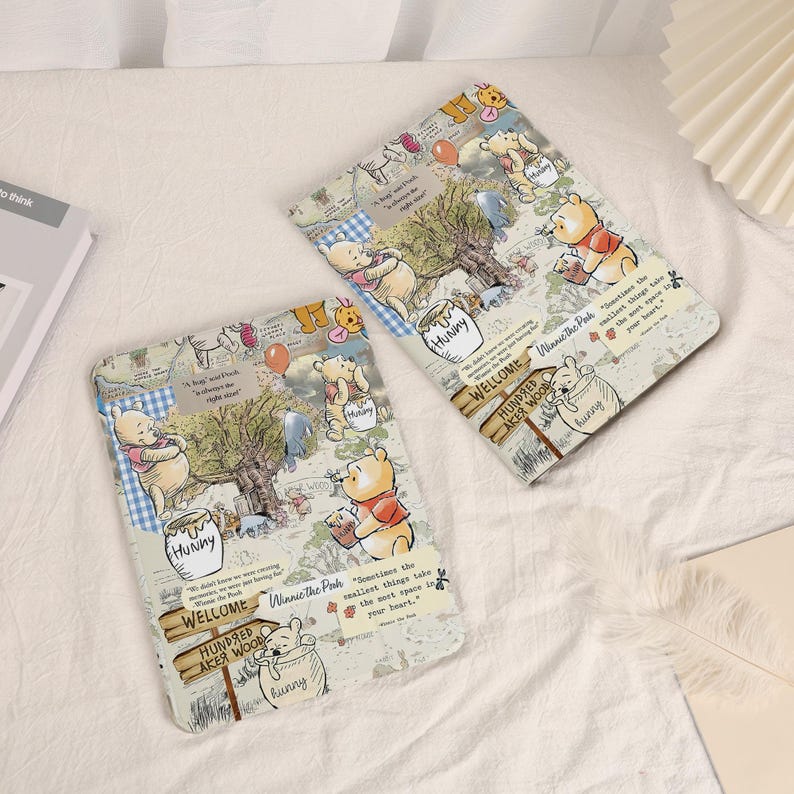 May include: Two rectangular cases featuring a Winnie the Pooh design. The cases have a collage-style pattern with illustrations of Pooh, trees, and text. The text includes "Hunny", "Welcome", and "Hundred Acre Wood". The cases are on a white surface.