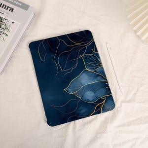 May include: A navy blue tablet case with a gold floral design. The case has a watercolor effect with gold outlines of leaves and a large flower. A white stylus rests beside the case on a white surface.