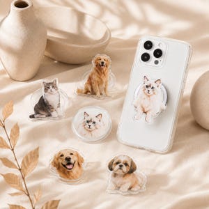 Peut inclure: Une collection de supports de téléphone en acrylique transparent présentant des portraits imprimés de diverses races de chiens et de chats, dont un golden retriever, un shih tzu et un chat blanc, fixés au dos d'une coque de smartphone.