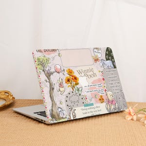 Puede incluir: Un portátil con una funda decorativa con personajes de Winnie the Pooh. La funda tiene un fondo rosa con ilustraciones de Pooh, Piglet, Eeyore y Tigger, junto con girasoles y texto. El portátil está sobre una superficie beige.