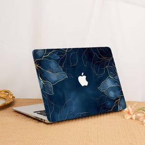 Könnte beinhalten: Ein Laptop mit einer dunkelblauen Schutzhülle, die mit einem goldenen Linienmuster aus Blumen verziert ist. Die Hülle hat eine glatte Oberfläche im Aquarellstil mit tiefblauen Farbtönen und einem weißen Apple-Logo in der Mitte.