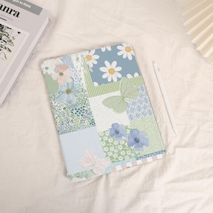 Può includere: Una custodia per tablet con motivi floreali con un design patchwork con margherite, farfalle e vari motivi verdi, blu e bianchi. Uno stilo bianco è appoggiato accanto alla custodia.