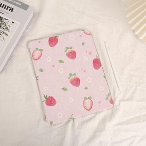 Könnte beinhalten: Eine rosa Tablet-Hülle mit einem sich wiederholenden Muster aus Erdbeeren, weißen Blumen und grünen Blättern. Ein weißer Stift liegt neben der Hülle. Die Hülle liegt auf einer weißen Oberfläche.