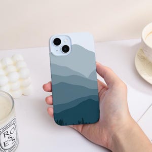 Op de afbeelding: Een telefoonhoesje met een berglandschapontwerp. De hoes heeft een verloop van blauwe en grijze tinten, met een silhouet van bomen aan de onderkant. De telefoonhoes wordt in een hand gehouden.
