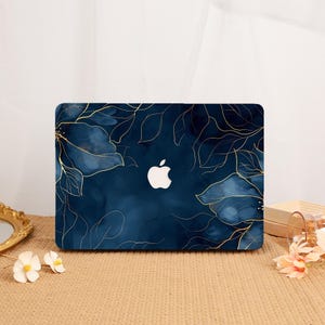 May include: A dark navy blue laptop cover featuring a watercolor-style floral pattern with delicate gold-colored line art outlines of leaves and petals. The center of the cover displays a cutout for the white Apple logo, creating a contrast.