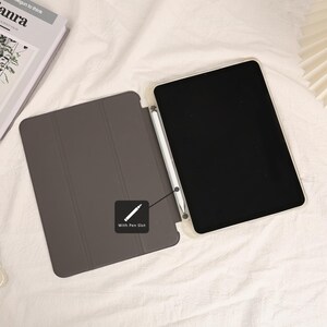 May include: A gray tablet case with a pen slot, open to reveal a black screen. A white stylus rests inside the case. The case is on a white surface. The text "With Pen Slot" is on the case.