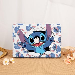 May include: A rectangular laptop skin featuring a repeating pattern of the blue character Stitch on a white background. In the centre, a large illustration shows Stitch appearing to break through the surface with arms outstretched and mouth open.