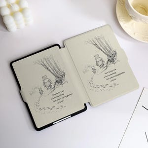 May include: A black e-reader case featuring a light beige interior with a black and white line drawing of Winnie the Pooh and Piglet walking away. The text reads, How lucky I am to have something that makes saying goodbye so hard.