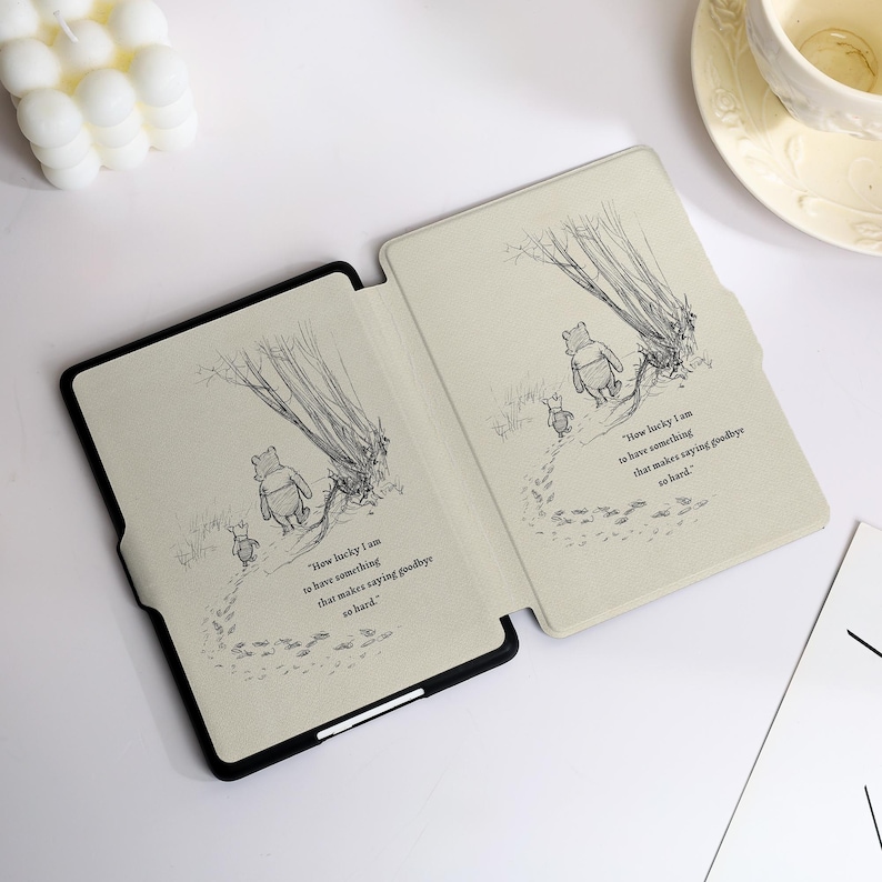 May include: A black e-reader case featuring a light beige interior with a black and white line drawing of Winnie the Pooh and Piglet walking away. The text reads, How lucky I am to have something that makes saying goodbye so hard.