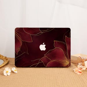 May include: A dark burgundy laptop hard shell case featuring a large, stylized floral pattern in thin, metallic gold line art. The design surrounds a central cutout that reveals the white Apple logo on the laptop lid underneath the cover.