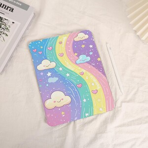 May include: A colorful tablet case featuring a rainbow design with clouds, hearts, and stars. The case has a pastel color scheme with a white stylus resting beside it. The case is on a white surface.