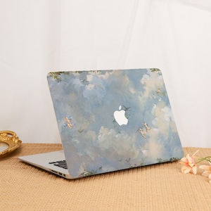 Puede incluir: Un ordenador portátil con un diseño pintado en azul y blanco con nubes y ángeles. El portátil tiene un logotipo de Apple blanco en la tapa.