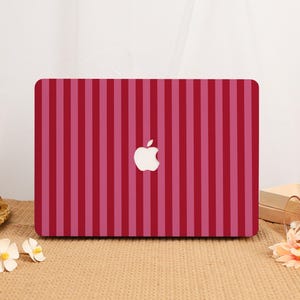 Puede incluir: Una funda para portátil con un patrón de rayas verticales en tonos alternos de rojo oscuro y rosa claro. El diseño cubre la tapa rectangular del dispositivo, que muestra un logotipo de Apple blanco recortado en el centro de las rayas.