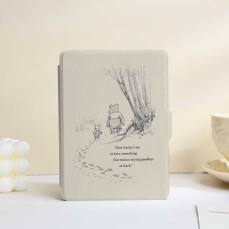 May include: A light beige e-reader case featuring a black line art illustration of Winnie the Pooh and Piglet walking away from the viewer. The text reads, "How lucky I am to have something that makes saying goodbye so hard." The case has a textured finish.