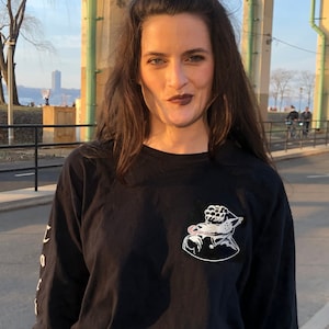 May include: A woman wearing a black long-sleeved shirt with a white graphic of a creature on it. She is wearing grey pants and a black belt. She is standing in front of a bridge with a city skyline in the background.
