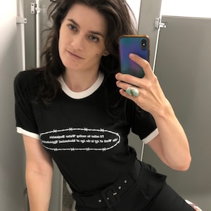 Könnte beinhalten: Eine Frau trägt ein schwarzes T-Shirt mit weißem Besatz und einem Stacheldraht-Design, auf dem steht "Versuch nicht, dich zu brüsten, es ist unmöglich zu sagen, ob du traurig bist oder nicht". Sie trägt eine schwarze Hose mit einem breiten Gürtel und hält ein Handy in der Hand.