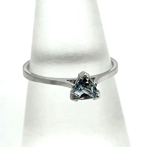 May include: A silver ring featuring a triangular, pale blue gemstone. The ring is presented on a clear, cone-shaped stand against a white backdrop. The gemstone is the central element, highlighting a minimalist design.