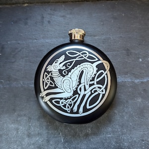 May include: Black metal flask with a silver Celtic knot design featuring a stylised wolf or dog.
