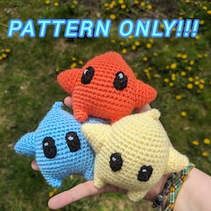 Puede incluir: Tres peluches de estrellas de ganchillo en diferentes colores: naranja, azul y amarillo. Cada peluche tiene dos ojos negros y una expresión simple y sonriente. Las peluches están en la mano de una persona.