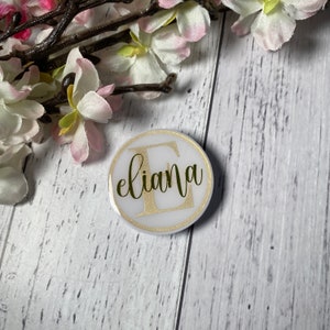 May include: White round sticker with a gold border and the name "eliana" written in cursive with a gold "E" in the centre.