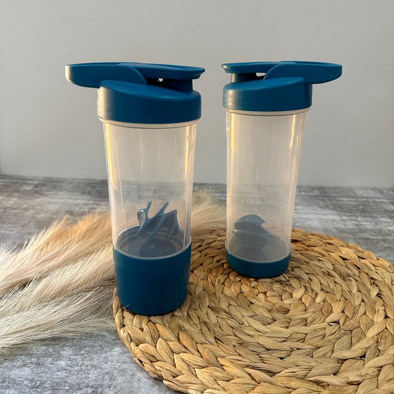 May include: Two blue and clear plastic shaker bottles with lids and mixing balls. The bottles are sitting on a woven straw mat.