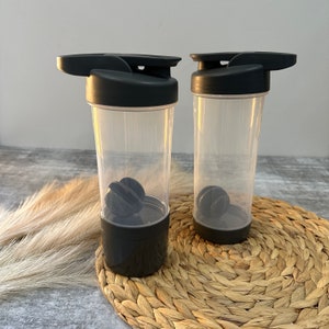 May include: Two black and clear plastic shaker bottles with black lids and a black mixing ball inside each bottle. The bottles are sitting on a woven straw mat.