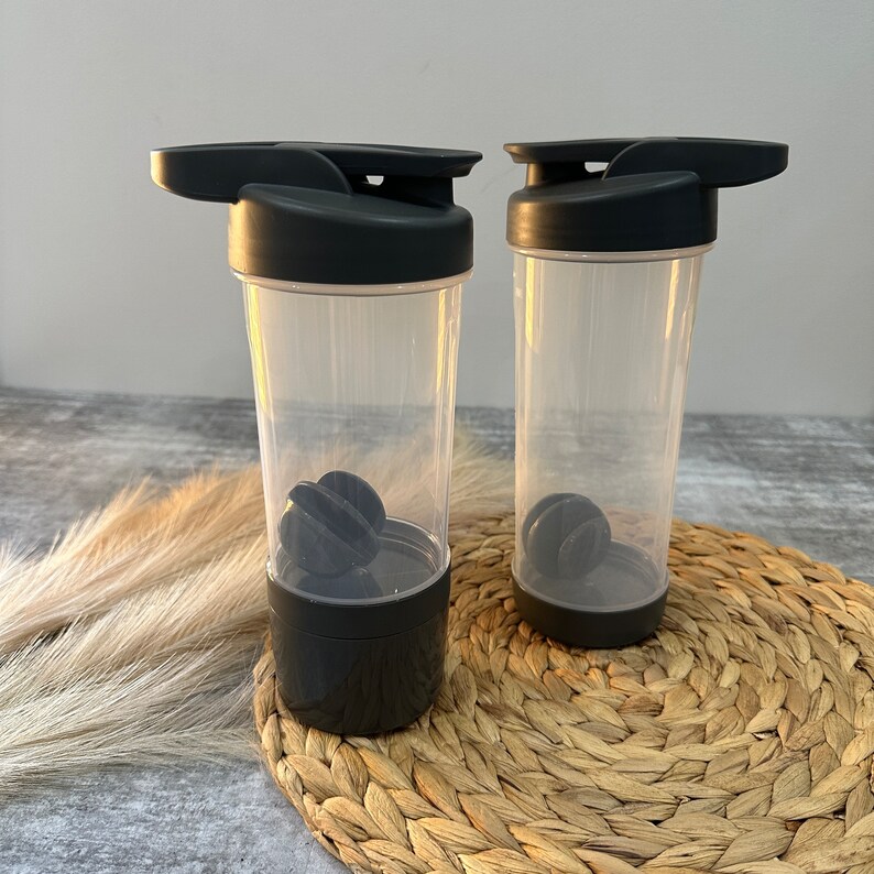 May include: Two black and clear plastic shaker bottles with black lids and a black mixing ball inside each bottle. The bottles are sitting on a woven straw mat.