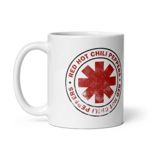 May include: White ceramic mug with the Red Hot Chili Peppers logo. The logo is a red asterisk inside a black circular border with the band's name. The mug has a curved handle and a glossy finish.