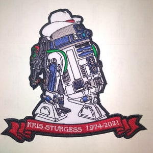 May include: An embroidered patch featuring a white, blue, and grey robot wearing a white fedora hat. Below the robot is a red banner with black borders containing the white embroidered text KRIS STURGESS 1974-2021. The design has a black outline.