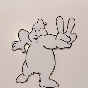 Puede incluir: Un parche bordado blanco que presenta un personaje de fantasma de dibujos animados con un cuerpo redondeado y un sombrero pequeño. El fantasma tiene una expresión sonriente y hace un signo de paz con una mano, delineado en hilo negro.