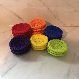 May include: A set of six crocheted coasters in rainbow colors: red, orange, yellow, green, blue, and purple. The coasters are stacked in a pyramid shape on a white marble surface.