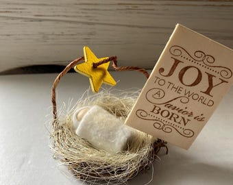 May include: A miniature nativity scene featuring a small wooden figure wrapped in white fabric resting in a straw nest. A yellow wooden star hangs from a brown wire arch. A rectangular wooden sign displays the text JOY TO THE WORLD A Savior is BORN.
