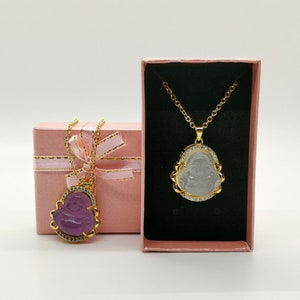 May include: Two gold necklaces with pendants in the shape of a Buddha. One pendant is clear and the other is purple. Both pendants are set with small clear stones. The necklaces are in pink gift boxes.