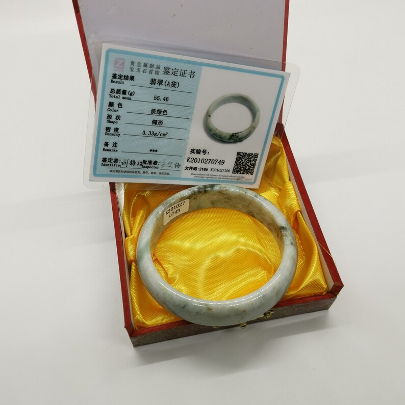 K&ouml;nnte beinhalten: Ein Jadeit-Bangle-Armband in hellgr&uuml;ner Farbe mit dunkelgr&uuml;nen Einschl&uuml;ssen. Das Armband befindet sich in einer roten und gelben Schachtel mit einem Echtheitszertifikat. Das Zertifikat besagt, dass das Armband 55,46 Gramm wiegt, hellgr&uuml;n ist und eine Dichte von 3,33 g/cm&sup2; hat. Die Zertifikatsnummer lautet K2010270749.