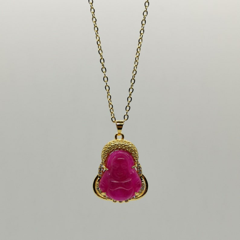 May include: A gold chain necklace with a pink jade pendant featuring a carved image of a Buddha. The pendant is surrounded by small clear stones.