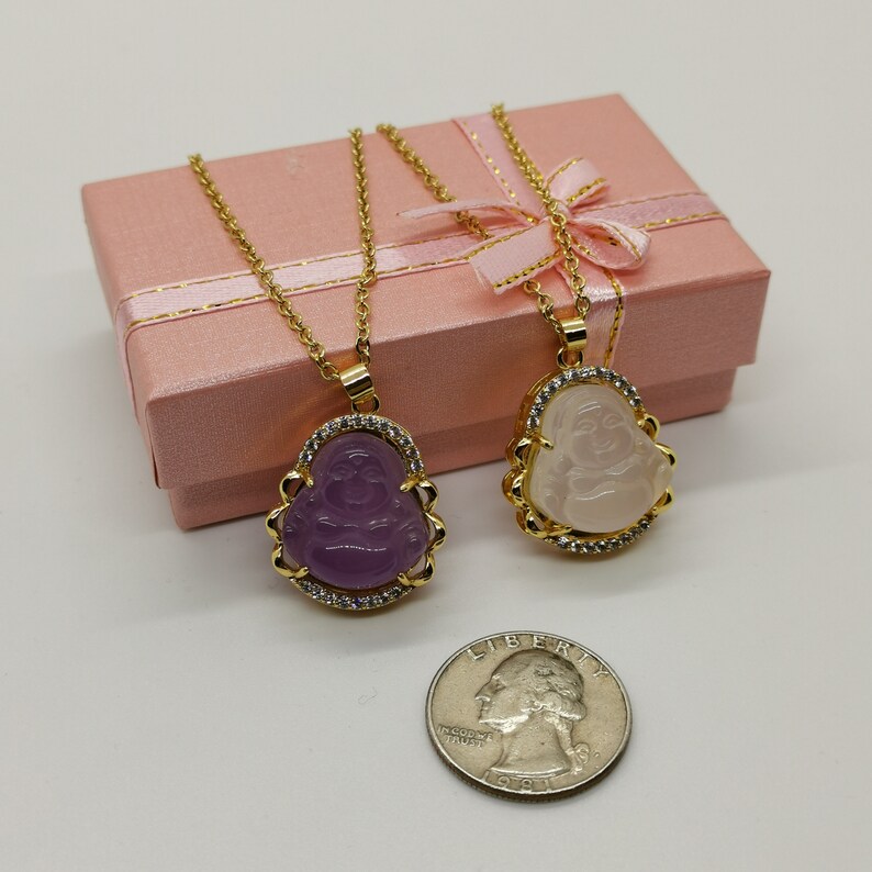 May include: Two gold necklaces with a  Laughing Buddha pendant. One pendant is purple and the other is white. Both pendants are surrounded by clear rhinestones.