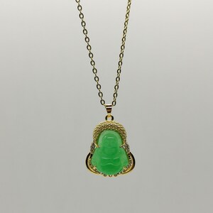 May include: A gold chain necklace with a green jade pendant featuring a Buddha figure. The pendant is surrounded by small clear stones.