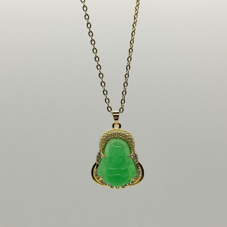 May include: A gold chain necklace with a green jade pendant featuring a Buddha figure. The pendant is surrounded by small clear stones.