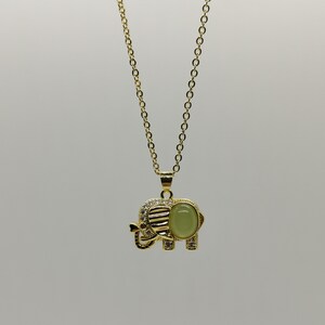 May include: A gold chain necklace with an elephant pendant. The elephant is decorated with clear rhinestones and has a green gemstone in the center.